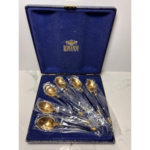 Puede incluir: Un juego de seis cucharas doradas con mangos negros, presentado en una caja forrada de terciopelo azul. La caja tiene un cierre dorado y la palabra "ROMANOV" impresa en dorado en el interior de la tapa. Las cucharas están envueltas individualmente en plástico transparente.