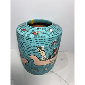 May include: A turquoise ceramic vase with a hand-painted design depicting a boat with people and sea creatures. The vase has a cylindrical shape with a wide opening and a wavy pattern. The artwork features red, white, and brown colours.