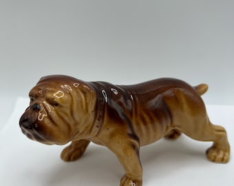 May include: A brown ceramic bulldog figurine with a wrinkled face and a collar. The figurine is standing on a white surface.