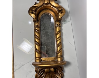 May include: Ornate gold-colored wall mirror with a rectangular shape and a small shelf at the bottom. The mirror has a decorative, scrolled top and a shell-like design at the base. The frame features a textured, column-like design.