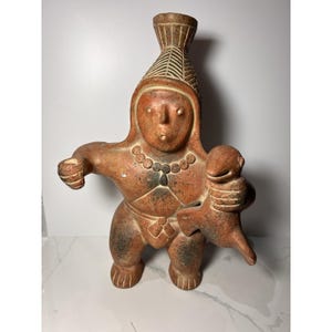 May include: A terracotta-colored clay sculpture of a figure wearing a tall, textured headdress and a beaded necklace. The figure holds a small animal in one arm and has one arm extended to the side. The surface features dark, mottled patches.