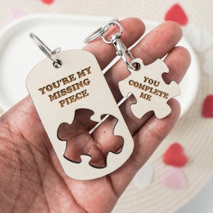 Puede incluir: Dos llaveros de madera con forma de piezas de rompecabezas. Un llavero dice "You're my missing piece" y el otro dice "You complete me".