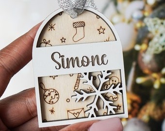 May include: White wooden Christmas ornament with a snowflake cutout and the name "Simone" written on it. The ornament is decorated with a variety of Christmas-themed illustrations.
