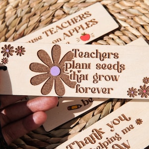 May include: Wooden bookmark with a brown daisy design and the text "Teachers plant seeds that grow forever". The bookmark has a black tassel.