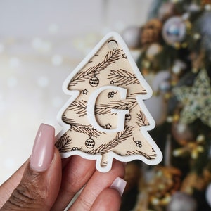 May include: White wooden Christmas tree bauble with a brown woodgrain pattern and the letter "G" in white. The bauble has a small hole at the top for hanging.