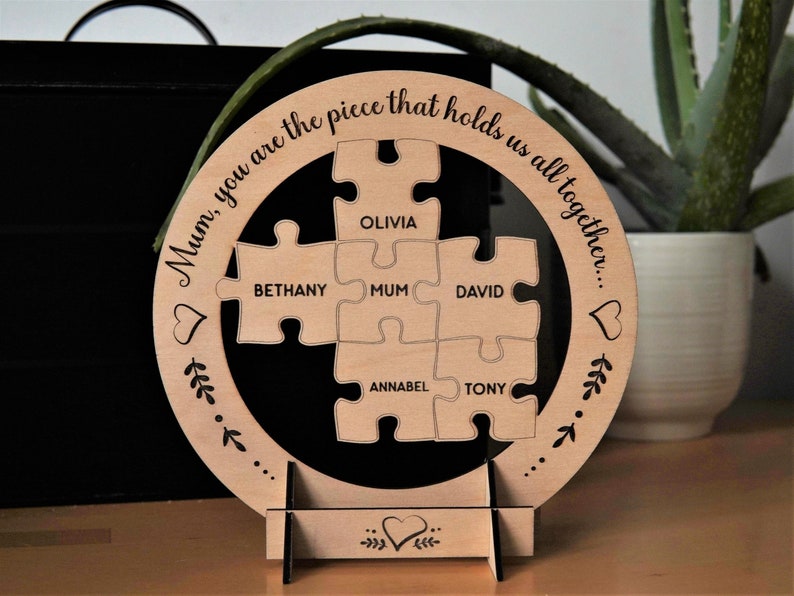 Może przedstawiać: Drewniane koło z wzorem układanki. Tekst "Mum, you are the piece that holds us all together..." jest wygrawerowany wok&oacute;ł krawędzi. Elementy układanki są oznaczone nazwami: Olivia, Bethany, Mum, David, Annabel i Tony.