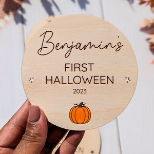 Puede incluir: Un letrero redondo de madera con el texto "Benjamin's First Halloween 2023" y una peque&ntilde;a ilustraci&oacute;n de una calabaza naranja.