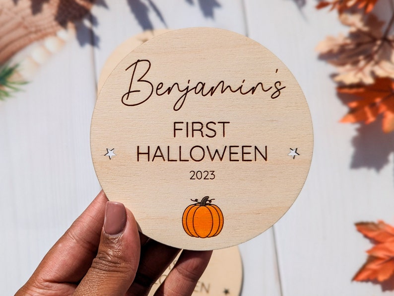 Puede incluir: Un letrero redondo de madera con el texto "Benjamin's First Halloween 2023" y una peque&ntilde;a ilustraci&oacute;n de una calabaza naranja.