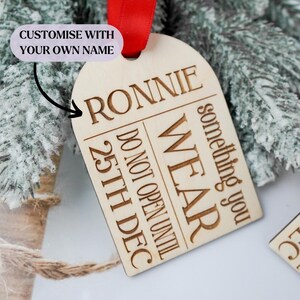 May include: A wooden Christmas ornament with the name "RONNIE" engraved at the top. The ornament has the text "WEAR something you DO NOT OPEN UNTIL 25TH DEC". A red ribbon is attached for hanging. The text "CUSTOMISE WITH YOUR OWN NAME" is also present.