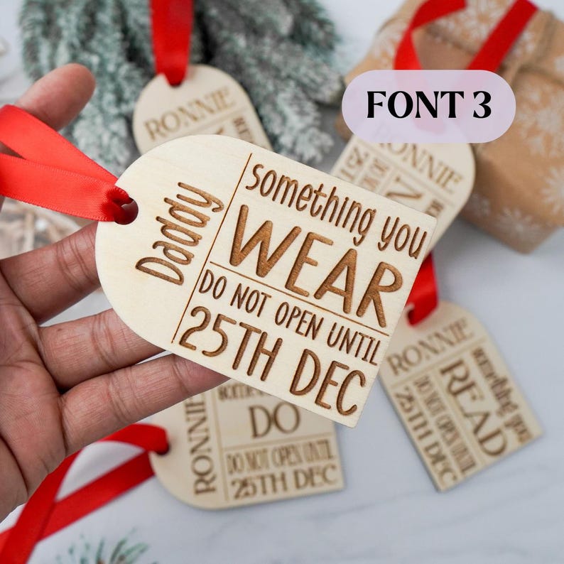May include: Wooden gift tags with red ribbon. One tag reads "Daddy" and "something you WEAR DO NOT OPEN UNTIL 25TH DEC." Other tags say "RONNIE" and "DO NOT OPEN UNTIL 25TH DEC." The tags are for Christmas gifts.