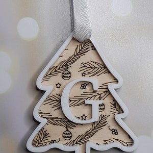 May include: White wooden Christmas tree ornament with a brown woodgrain pattern and the letter "G" cut out in the center. The ornament has a white ribbon for hanging.