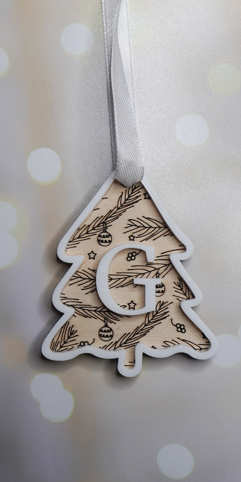 May include: White wooden Christmas tree ornament with a brown woodgrain pattern and the letter "G" cut out in the center. The ornament has a white ribbon for hanging.