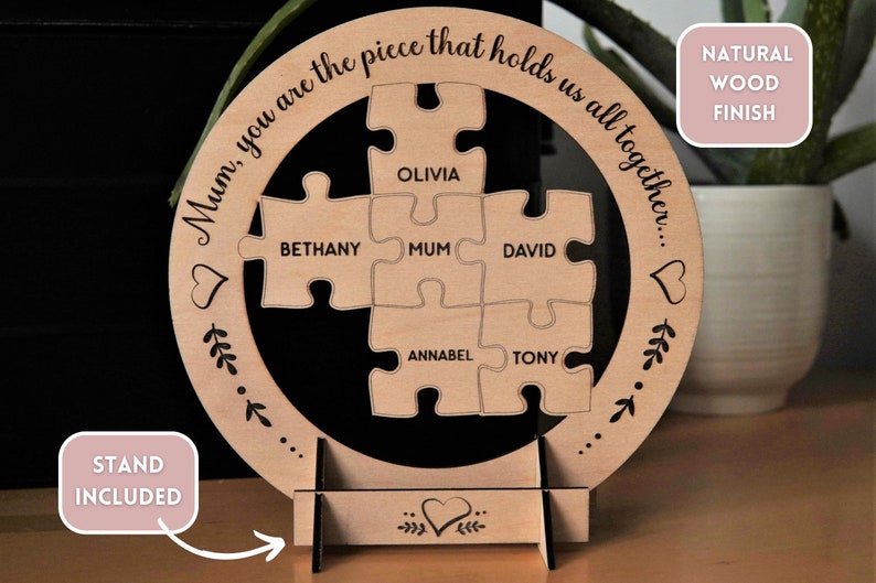 Może przedstawiać: Okrągła drewniana tabliczka z wzorem układanki. Elementy układanki są oznaczone nazwami, w tym "Mama", "Olivia", "Bethany", "David", "Annabel" i "Tony". Tabliczka zawiera tekst "Mamo, jesteś częścią, kt&oacute;ra nas wszystkich łączy..." i "Naturalne wykończenie drewna". W zestawie znajduje się stojak.