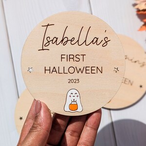 Puede incluir: Un letrero redondo de madera con el texto "Isabella's First Halloween 2023" y un fantasma de dibujos animados sosteniendo una calabaza. El letrero tiene dos peque&ntilde;as estrellas blancas a los lados.