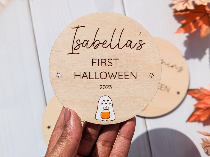 Puede incluir: Un letrero redondo de madera con el texto "Isabella's First Halloween 2023" y un fantasma de dibujos animados sosteniendo una calabaza. El letrero tiene dos peque&ntilde;as estrellas blancas a los lados.
