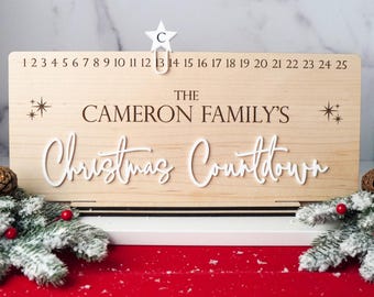 Può includere: Un calendario dell'avvento in legno con il testo "The Cameron Family's Christmas Countdown" e i numeri da 1 a 25. Una stella bianca con la lettera "C" è in cima al calendario.
