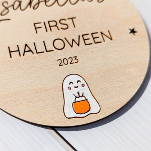 Puede incluir: Un letrero redondo de madera con el texto "Isabella First Halloween 2023" y un fantasma de dibujos animados sosteniendo un cubo de dulces.