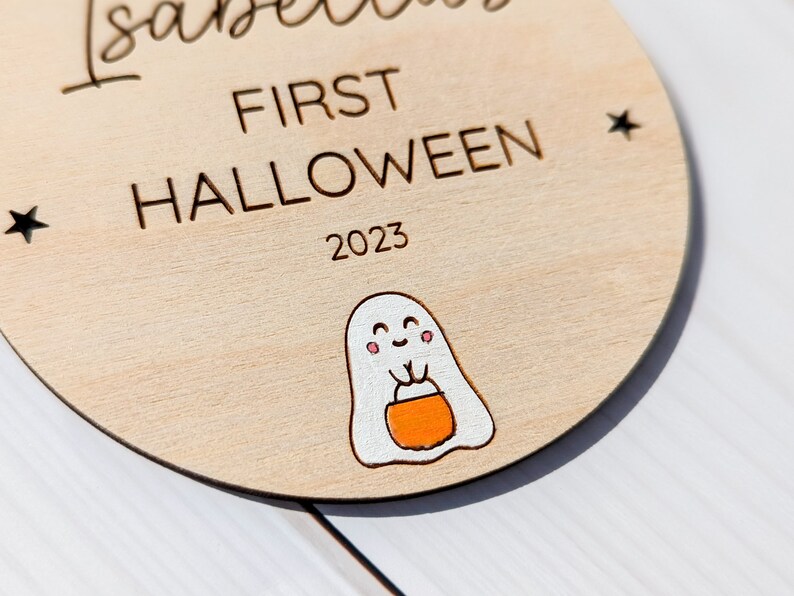 Puede incluir: Un letrero redondo de madera con el texto "Isabella First Halloween 2023" y un fantasma de dibujos animados sosteniendo un cubo de dulces.