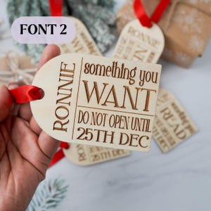 May include: Wooden gift tags with red ribbon. The tags have the name "RONNIE" and the text "something you WANT DO NOT OPEN UNTIL 25TH DEC". Other tags are visible in the background.