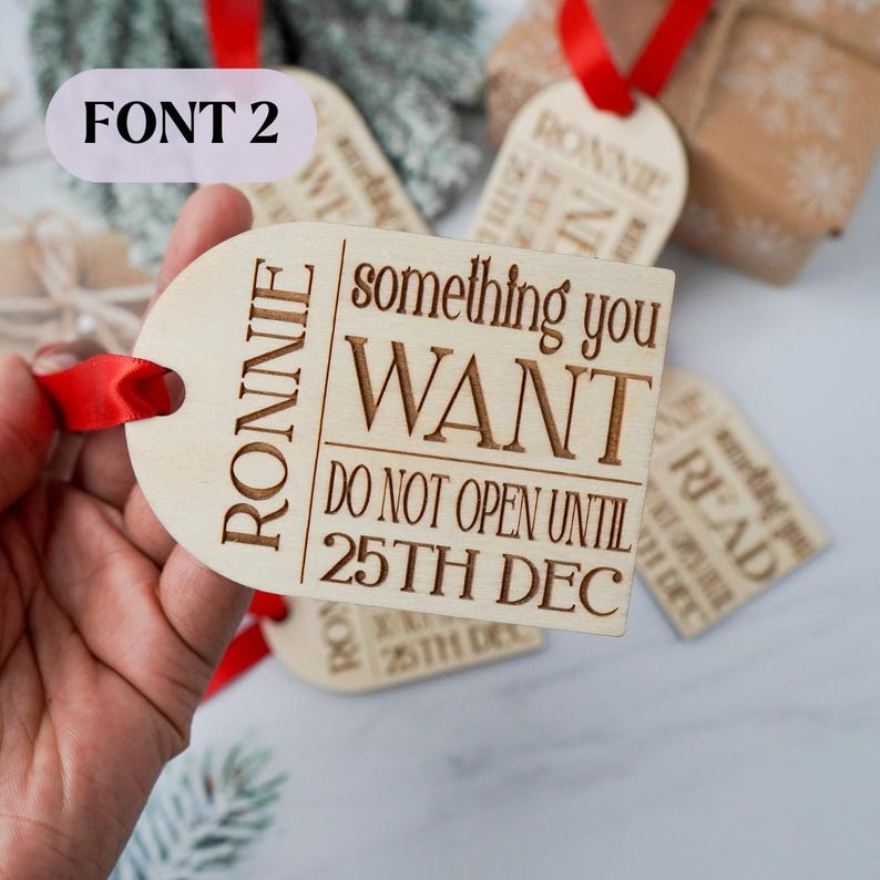 May include: Wooden gift tags with red ribbon. The tags have the name "RONNIE" and the text "something you WANT DO NOT OPEN UNTIL 25TH DEC". Other tags are visible in the background.