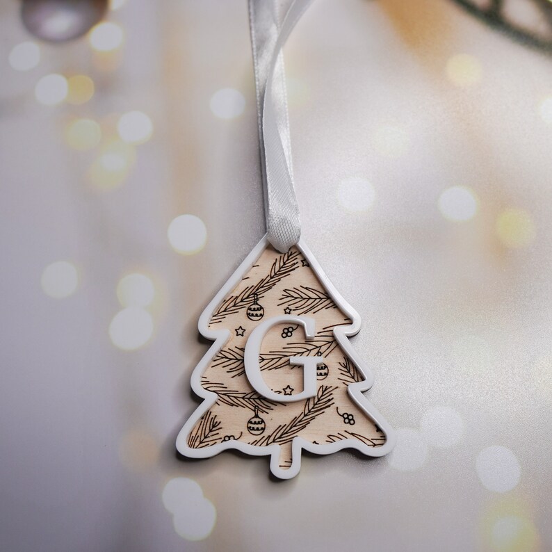 May include: White wooden Christmas tree ornament with a white letter G in the center. The ornament has a white ribbon for hanging and a brown and white pattern of pine branches and ornaments.