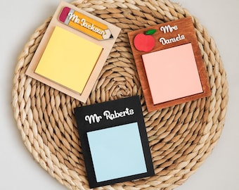 May include: Three rectangular wooden sticky note holders with personalized names. One features a yellow pencil and 'Ms Jackson', one a red apple and 'Mrs Daniels', and one is black with 'Mr Roberts'. Each holds a square pad of paper.
