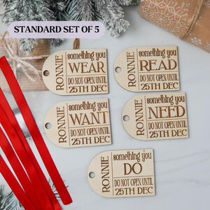 May include: A set of five wooden gift tags with the name "RONNIE" and the phrases "WEAR", "READ", "WANT", "NEED", and "DO". Each tag includes "DO NOT OPEN UNTIL 25TH DEC". Red ribbon is visible.