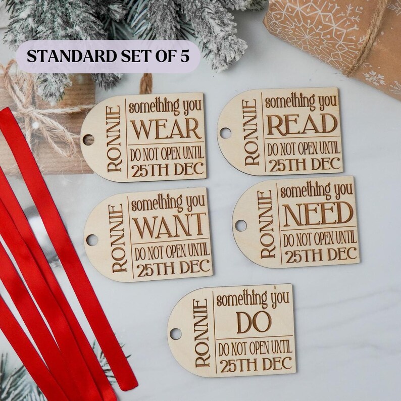 May include: A set of five wooden gift tags with the name "RONNIE" and the phrases "WEAR", "READ", "WANT", "NEED", and "DO". Each tag includes "DO NOT OPEN UNTIL 25TH DEC". Red ribbon is visible.