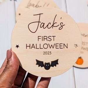 Puede incluir: Un letrero redondo de madera con el texto "Jack's First Halloween 2023" y una silueta de murci&eacute;lago negro. El letrero est&aacute; decorado con dos peque&ntilde;as estrellas.