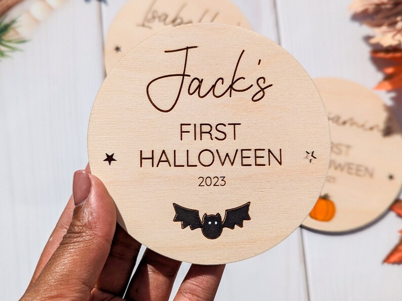 Puede incluir: Un letrero redondo de madera con el texto "Jack's First Halloween 2023" y una silueta de murci&eacute;lago negro. El letrero est&aacute; decorado con dos peque&ntilde;as estrellas.