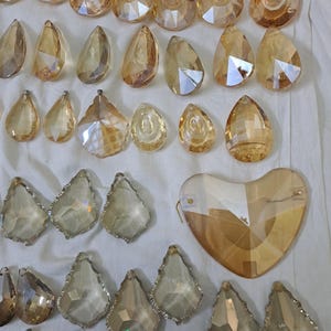 May include: An assortment of amber-colored crystal chandelier pendants in various shapes, including teardrops, faceted gems, and a heart. The crystals are arranged on a white surface, reflecting light and creating a sparkling effect. Ideal for home decor and lighting projects.