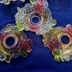 May include: Clear glass decorative pieces with rose and leaf designs. The pieces have a central hole and are colored with red, yellow, and green hues. The glass pieces are arranged on a dark blue surface.