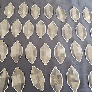 May include: A collection of clear, faceted crystal chandelier pendants arranged on a dark blue surface. Each pendant has a unique, elongated shape with multiple facets, reflecting light. These decorative crystals are ideal for home decor and craft projects.