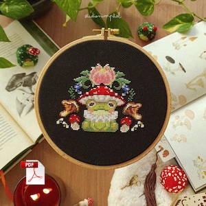 Pode incluir: Um bastidor de bordado circular de madeira contendo um design em ponto cruz sobre tecido preto. O design apresenta um sapo verde usando um chapéu de cogumelo vermelho e branco, cercado por cogumelos menores, folhas verdes e uma flor rosa.