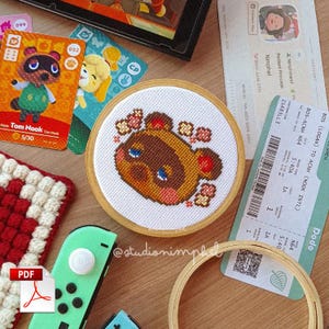 Pode incluir: Um bastidor de bordado circular de madeira contendo um padrão de ponto cruz da personagem Tom Nook de Animal Crossing. O design apresenta um rosto de guaxinim castanho com olhos azuis, bochechas rosadas e pequenos detalhes florais em tecido branco.