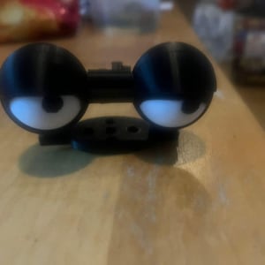 May include: A 3D-printed black and white object, designed to look like cartoon eyes. The eyes are spherical, with black pupils and white irises. It has a connecting bar and a circular base, possibly a component or decorative piece.