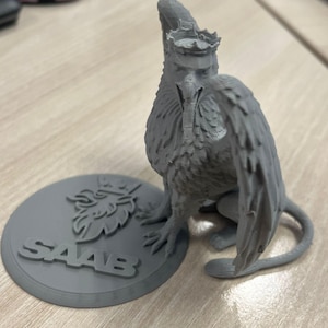 Könnte beinhalten: Eine graue, 3D-gedruckte Greifenfigur mit Krone, detaillierten Flügeln und einem gebogenen Schwanz. Der Greif steht neben einem runden Sockel mit dem Wort "SAAB" und einem Logo. Die Figur befindet sich auf einer hellen Oberfläche.