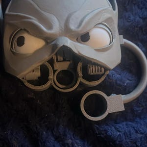 Könnte beinhalten: Ein grauer, 3D-gedruckter Becher mit einem stilisierten Gesicht, ausgeprägten Augenbrauenwülsten, großen weißen Augen mit schwarzen Pupillen und einem mechanisch wirkenden Unterteil. Der Becher hat einen geschwungenen Henkel und einen separaten Ring.