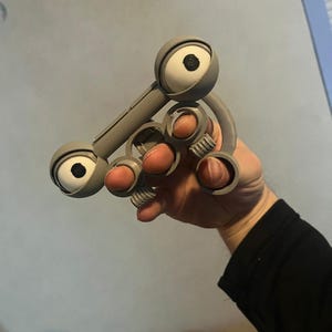 Puede incluir: Un juguete articulado gris impreso en 3D con dos grandes características redondas en forma de ojo y varios anillos del tamaño de un dedo. Los ojos tienen iris blancos y pupilas negras. El juguete se sostiene en una mano.
