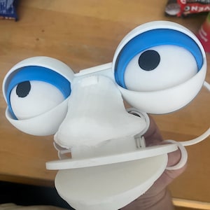 Puede incluir: Un rostro de juguete de plástico blanco con dos ojos grandes y redondos, párpados azules y pupilas negras. El juguete tiene una nariz blanca moldeada prominente entre los ojos y una pieza bucal blanca, plana y semicircular en la base.