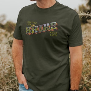 May include: A dark green t-shirt with a vintage-style graphic that says "Greetings from the Shire Middle Earth".