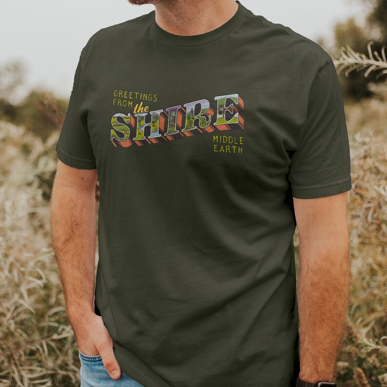 May include: A dark green t-shirt with a vintage-style graphic that says "Greetings from the Shire Middle Earth".