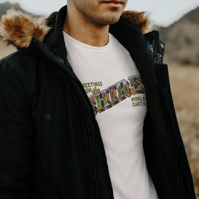May include: White t-shirt with a vintage postcard design featuring the text "Greetings from the Shire, Middle Earth".