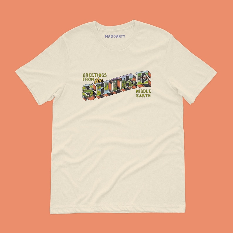 May include: A cream colored t-shirt with a vintage style graphic that says "Greetings from the Shire Middle Earth".