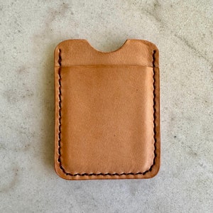 May include: A light brown leather card holder with a curved top opening. The edges are stitched with a darker thread, and the overall design is minimalist. The card holder is rectangular and appears to be handmade.