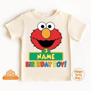 以下が含まれることがあります： ベージュのTシャツに、赤いエルモのキャラクターと、「3 NAME BIRTHDAY BOY！」と書かれた緑色の看板が描かれています