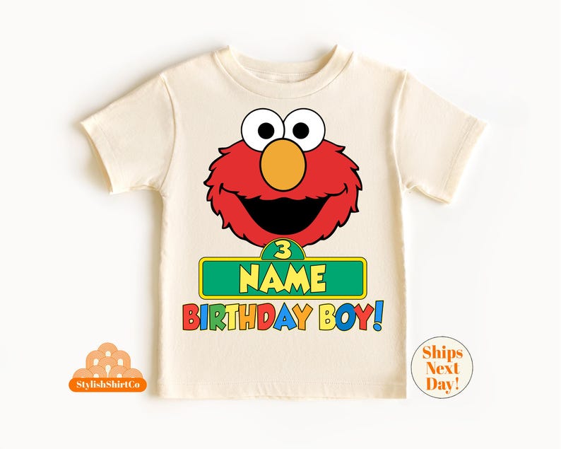 以下が含まれることがあります： ベージュのTシャツに、赤いエルモのキャラクターと、「3 NAME BIRTHDAY BOY！」と書かれた緑色の看板が描かれています