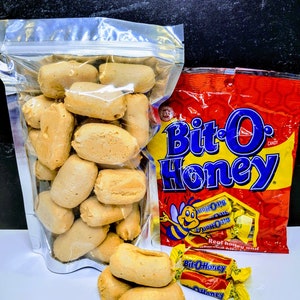 May include: A clear plastic bag filled with honey-flavored puffed wheat cereal snacks. Next to it is a red bag of Bit-O-Honey candy with a cartoon bee and the text "Bit-O-Honey" in yellow letters. There are also three Bit-O-Honey candies in the image.
