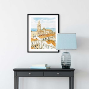 May include: A black framed watercolor painting of a cityscape with a tall tower and a domed building. The painting is hanging on a white wall above a black console table with two drawers. A blue lamp with a white shade is on the table.