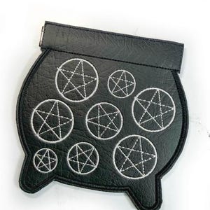 May include: A black faux leather patch shaped like a cauldron, featuring eight embroidered silver pentagrams of varying sizes arranged across the surface. The patch has a distinct top rim and two small feet at the bottom, with stitched edges.